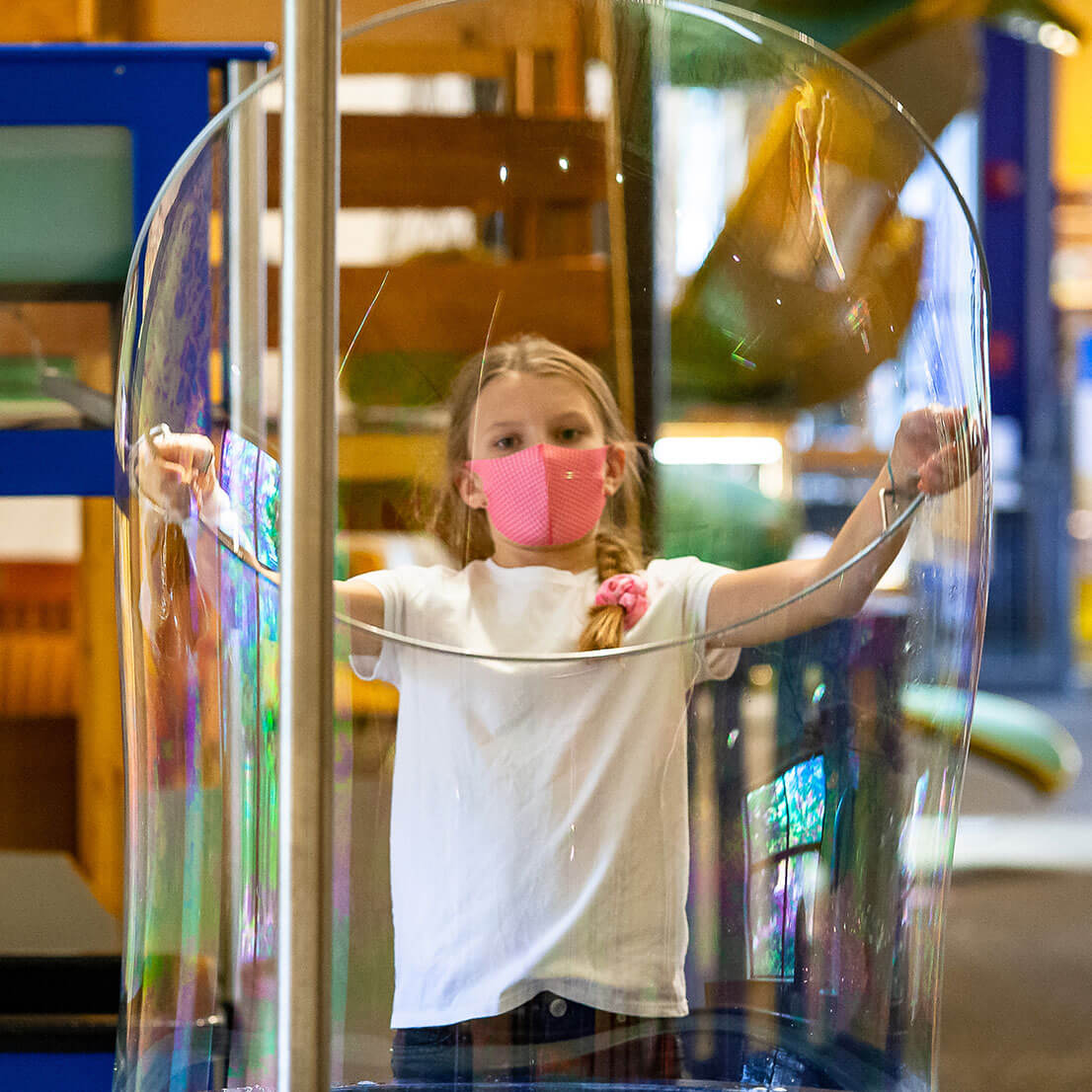 A young girl wearing a face mask is creating a giant bubble with a large ring in Wonderlab's bubble area