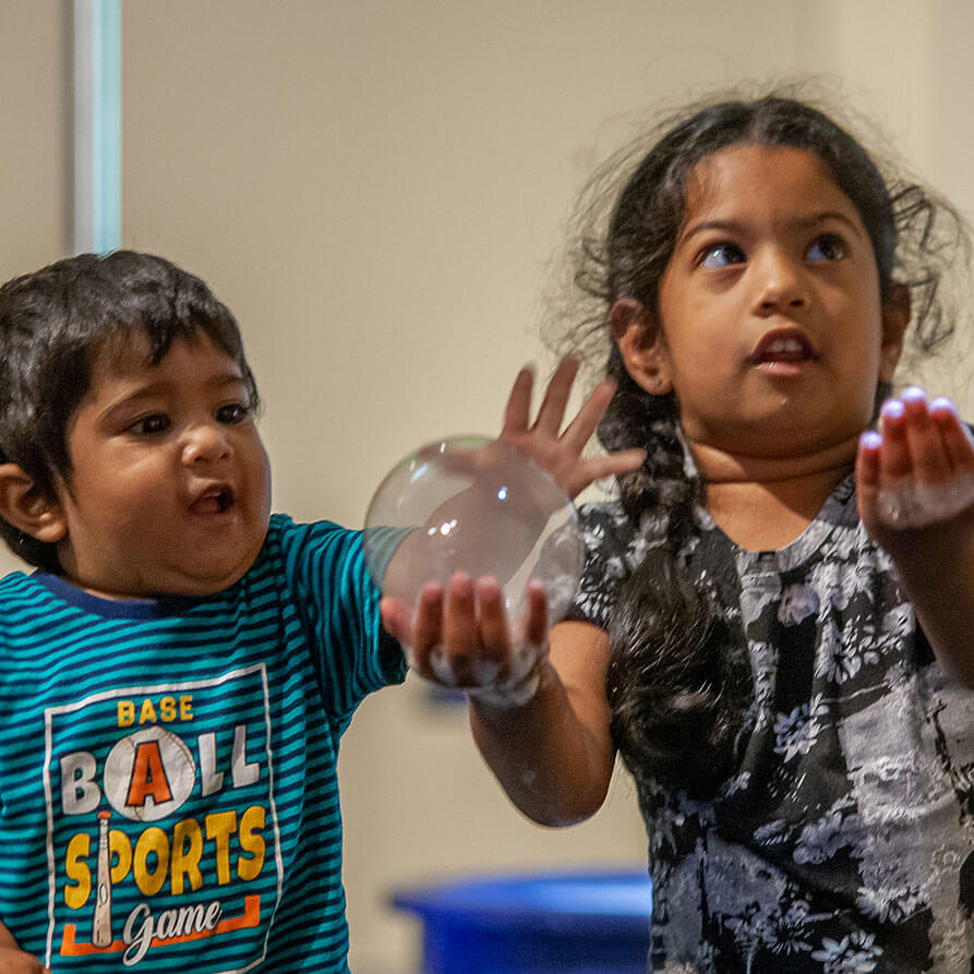 A child holds a large bubble in her had, while a younger child reaches for it
