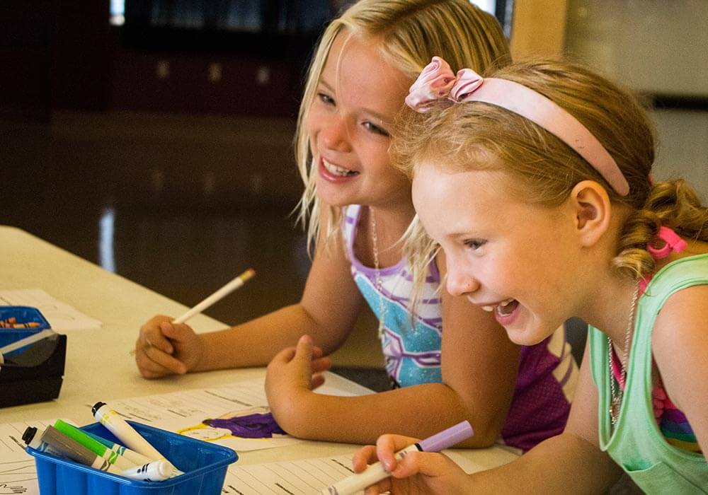 Two girls are writing on worksheets at a table, laughing, at Wonderlab in Bloomington