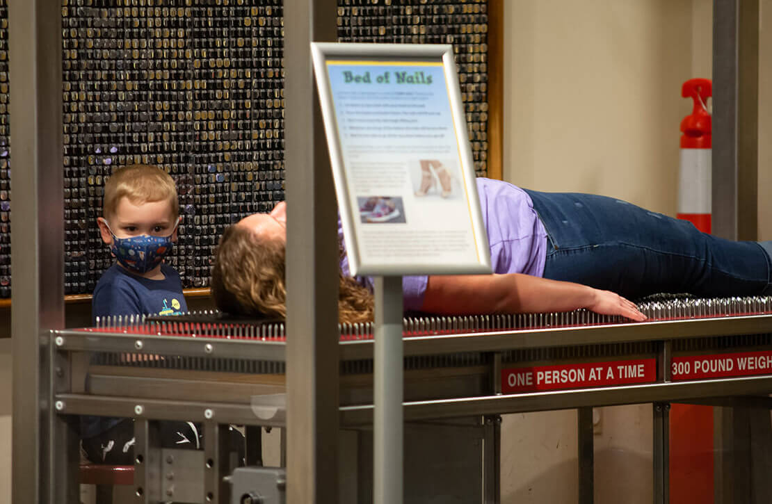 A child lays on the Bed of Nails exhibit at Wonderlab while a younger child looks on