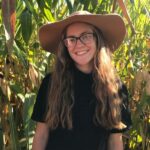 Photo of author Tavia Hedrick, a smiling, light skinned young woman with long hair standing in a forest wearing a wide brimmed hat and glasses.