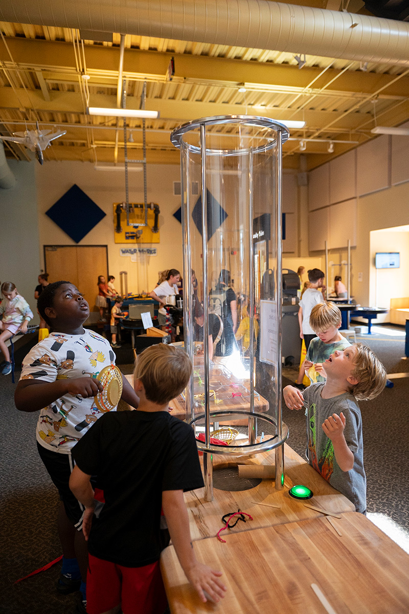 Four children gather around a translucent wind tunnel watching as a column of air makes things fly upward.