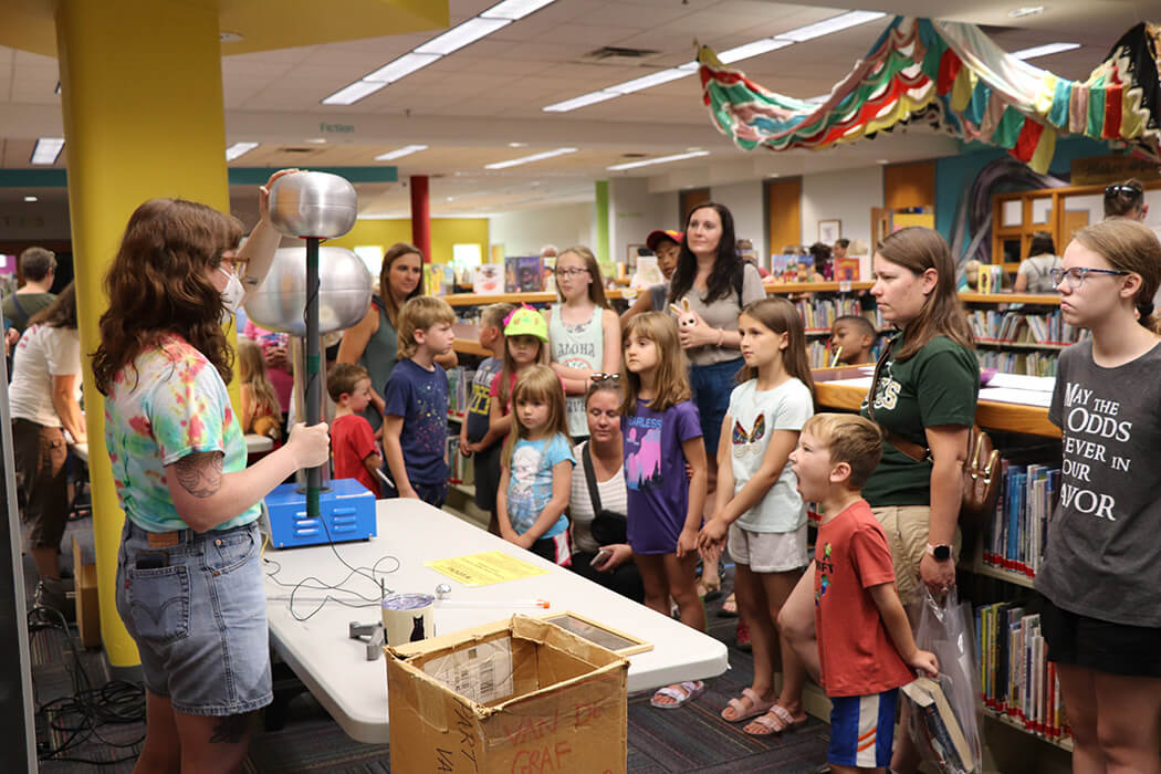 Several children of varying ages gather around to watch a Wonderlab science experiment in a library