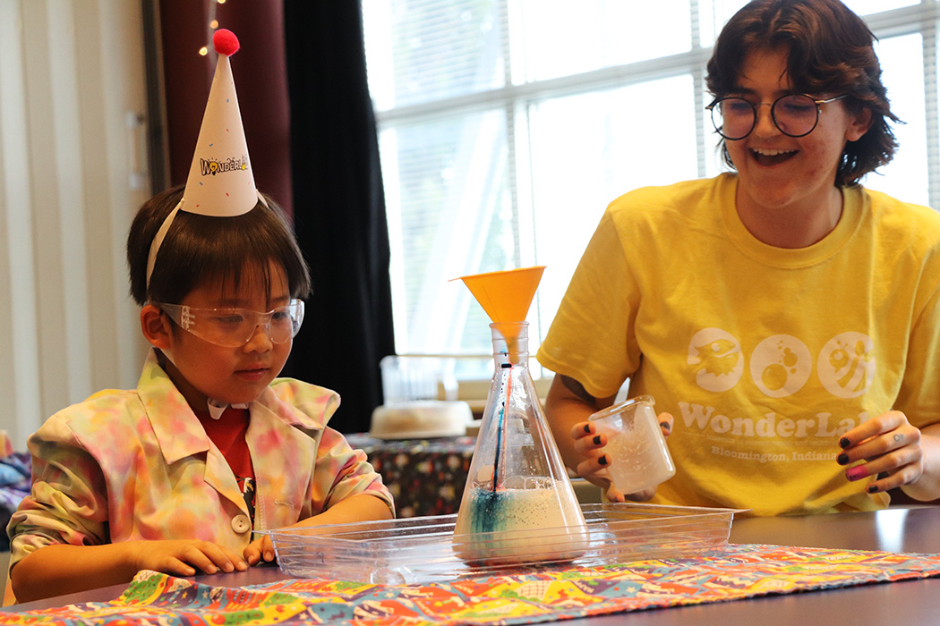 A young boy in a birthday party hat and a young woman wearing a WonderLab t-shirt. watches a chemical reaction in a conical flask.