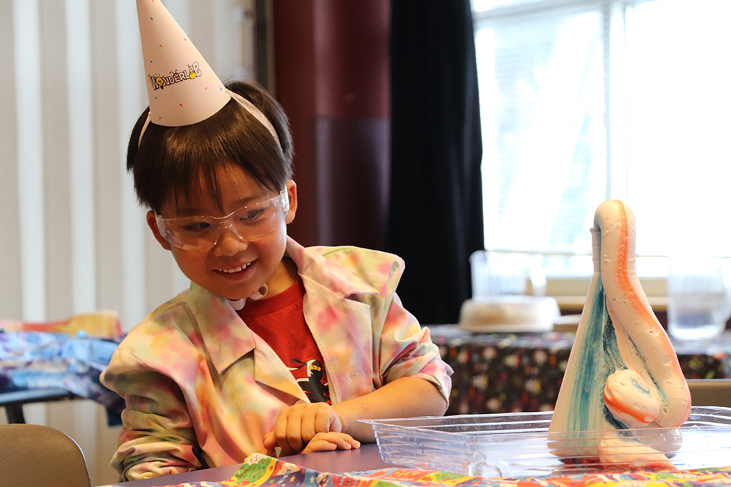 A young boy in a WonderLab birthday party hat watches a chemical reaction in a conical flask.