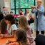 Middle aged woman leads a group of elementary aged children in a science experiment.