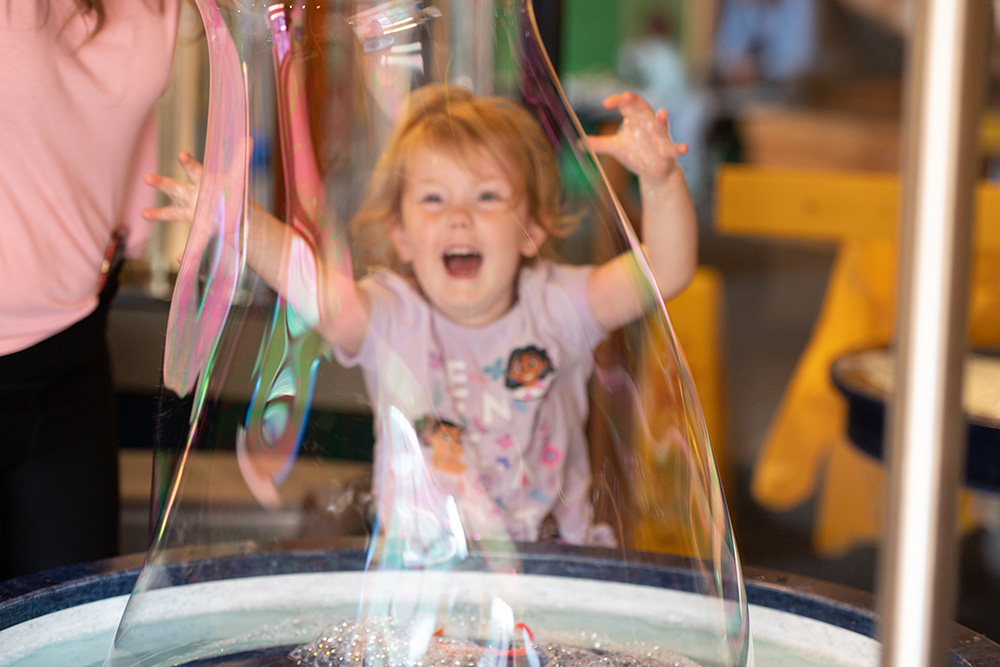 A toddler smiles with glee at a giant bubble being made in front of her at WonderLab Bloomington
