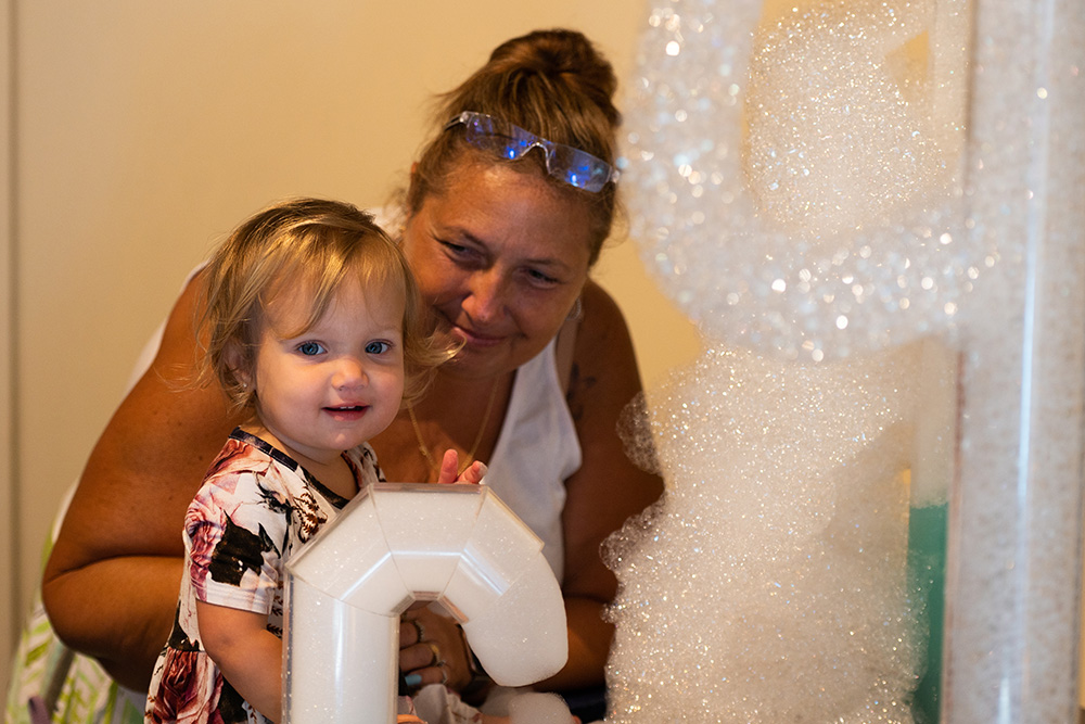 A small child with her caregiver at BubbleFest smiles as she looks at lots of foamy bubbles